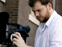 A man in a light striped shirt adjusts the settings on a large professional video camera.