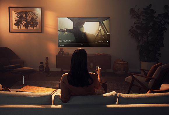 A person sits on a couch, facing a large television playing a film in a warm, dimly lit living room.