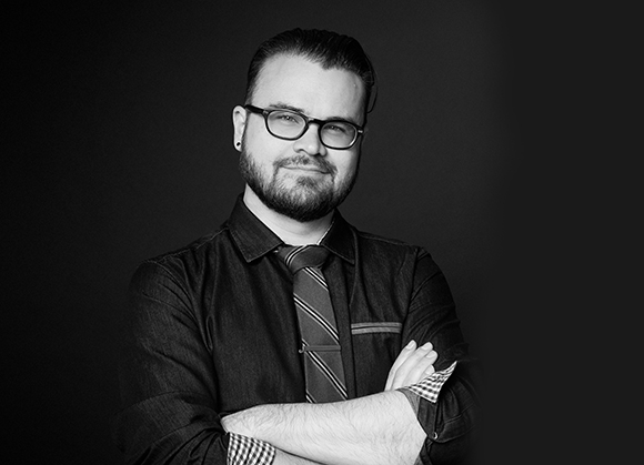 A bearded man wearing glasses, a dark button-down shirt, and a striped tie poses with arms crossed.