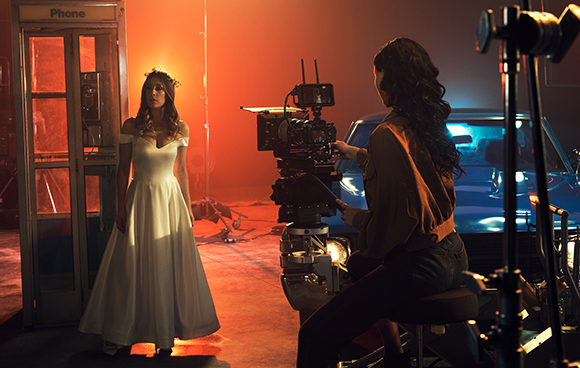 A woman in a white gown stands by a phone booth while a camera operator films her in warm, orange lighting.