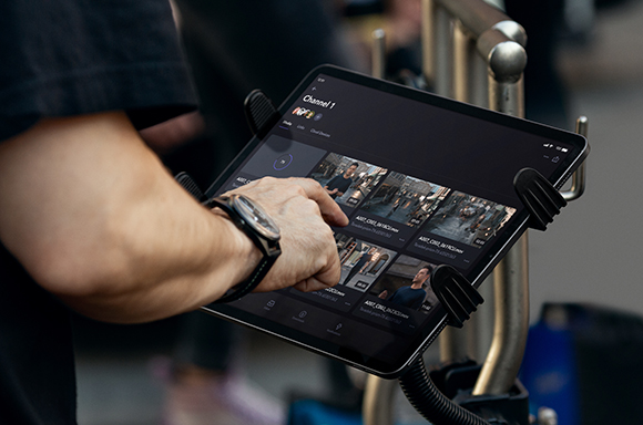 A person's hand touches a tablet screen displaying a video gallery interface, mounted on a professional stand.