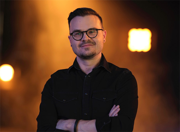 A man with short dark hair and glasses wearing a black button-down shirt, smiling with arms crossed.