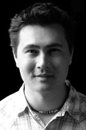 Black and white headshot of a man wearing a plaid shirt and a necklace against a dark background.