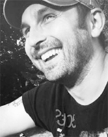 Black and white close-up of a smiling man with facial hair wearing a baseball cap.