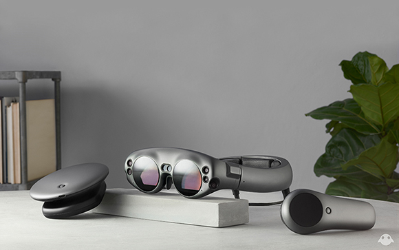 Dark grey augmented reality glasses, a small circular controller, and a charging case on a white table.