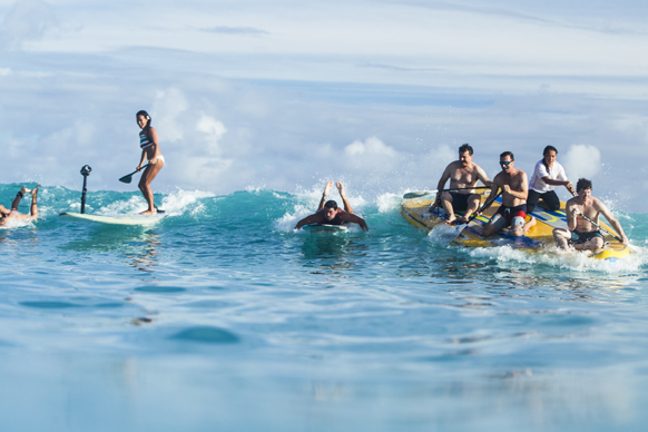 A woman paddleboards on a wave while others surf on longboards in the ocean.