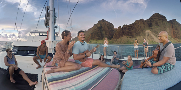 People sit and play music on a boat deck with large, green mountains in the background.