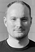Black and white headshot of a man with short hair and light facial stubble looking at the camera.