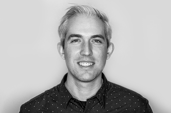 Black and white headshot of a man with light hair and a patterned button-down shirt smiling at the camera.