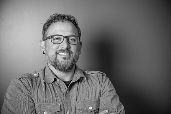 A smiling man with glasses and a beard wearing a button-down shirt against a plain background.