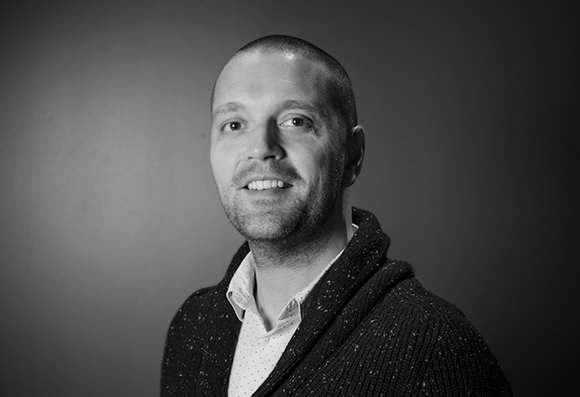 Black and white headshot of a man with short hair and light facial stubble, wearing a collared shirt and cardigan.