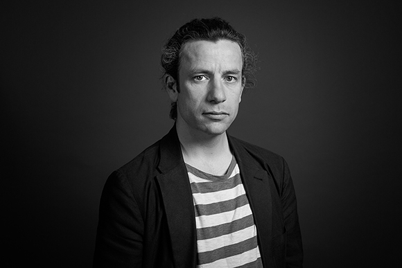 Black and white studio portrait of a man with curly hair wearing a dark blazer and a striped t-shirt.