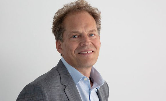 A man with curly light brown hair wearing a grey plaid blazer and a light blue button-down shirt.