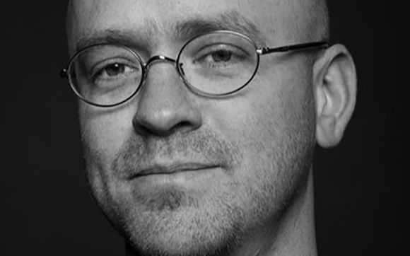 Black and white close-up portrait of a man with short hair wearing round-rimmed glasses.