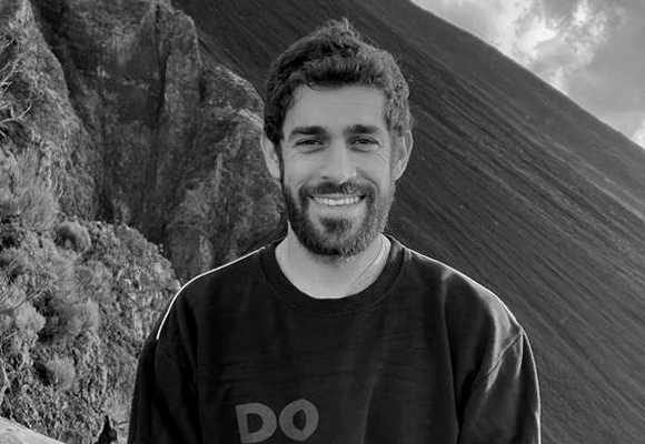 Black and white portrait of a smiling man with a beard standing before a steep, textured mountain slope.