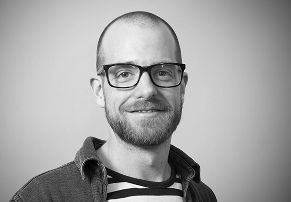 Black and white headshot of a man with short hair, glasses, and a beard wearing a striped shirt and denim jacket.
