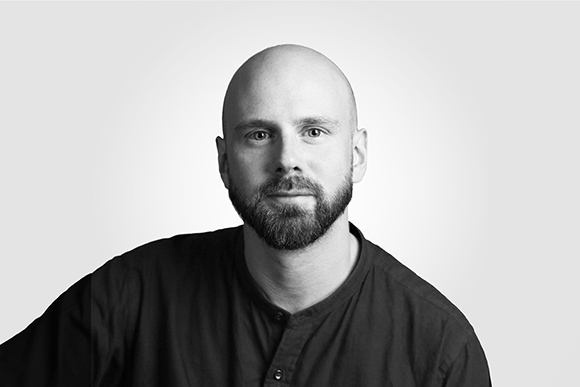 Black and white studio portrait of a bald man with a dark beard wearing a dark button-up shirt.