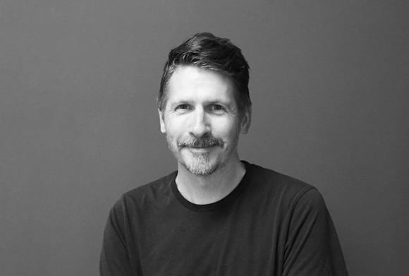 A black and white portrait of a man with short hair and a mustache smiling against a plain background.