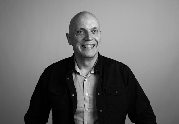 A smiling bald man wearing a dark open shirt over a light button-down shirt against a plain background.