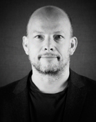 Black and white headshot of a bald man with a light beard wearing a dark jacket and t-shirt.