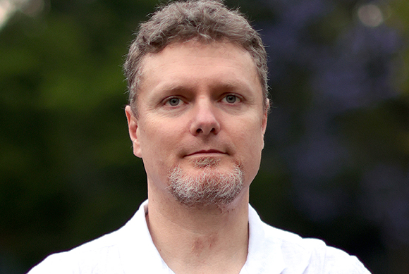 A man with short, wavy light brown hair and a greyish goatee wearing a white shirt.