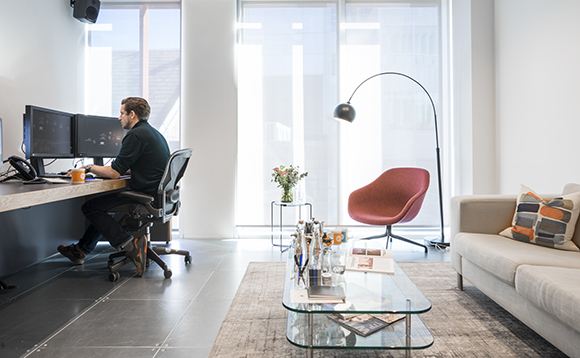 A man sits at a desk with dual monitors in a bright office with a sofa and glass coffee table.