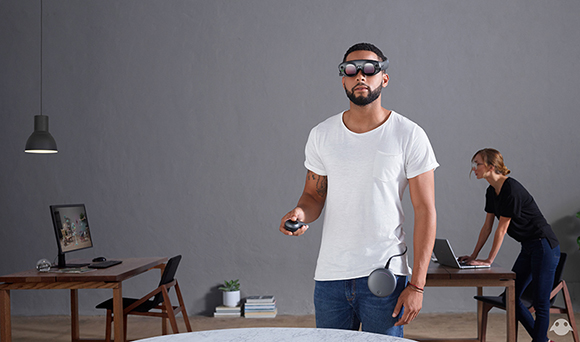 A man in a white t-shirt wears AR glasses and holds a controller in a modern office.