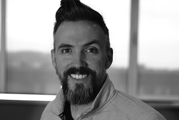 Black and white headshot of a man with a dark beard and styled hair, smiling at the camera.