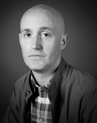 Black and white headshot of a bald man wearing a jacket and a plaid shirt.