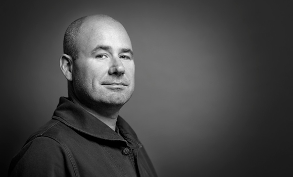Black and white portrait of a bald man wearing a dark jacket against a dark background.
