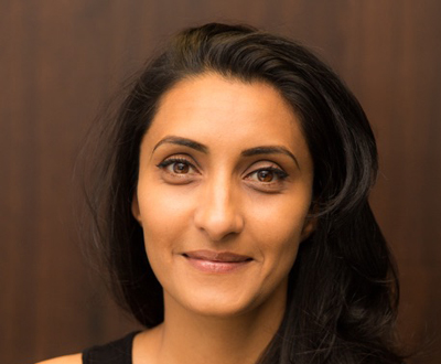 A woman with dark hair and brown eyes smiling against a dark brown background.