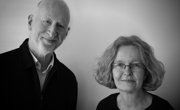 A black and white portrait of a bald man in a dark jacket and a woman with wavy hair and glasses.