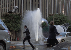 A man runs past a large water fountain and a massive silver sculpture in front of a tall building.