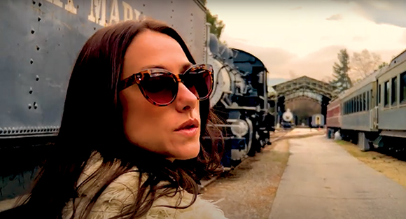 A woman wearing tortoiseshell sunglasses looks toward the camera in front of large blue steam locomotives.