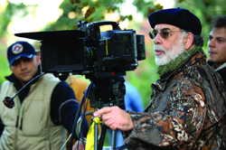A man wearing a black beret and camouflage jacket operates a large professional film camera outdoors.