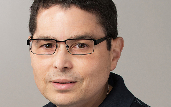 A man with short dark hair and black-rimmed glasses looking toward the camera against a gray background.
