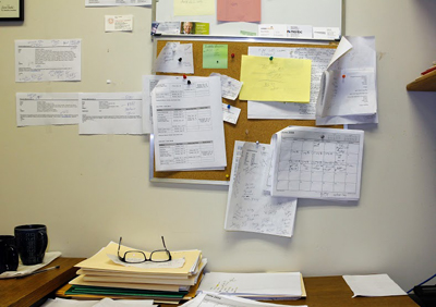 A corkboard with many pinned papers, notes, and calendars above a desk with stacks of files and glasses.