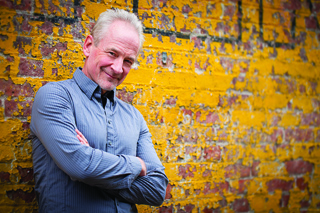A man with graying hair and a blue button-down shirt smiles with arms crossed against a yellow brick wall.