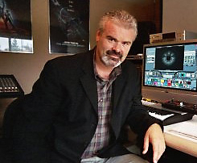 A man with graying hair and a beard wearing a dark blazer sits in a studio with monitors.