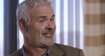 A smiling man with gray hair and a beard wearing a brown blazer.