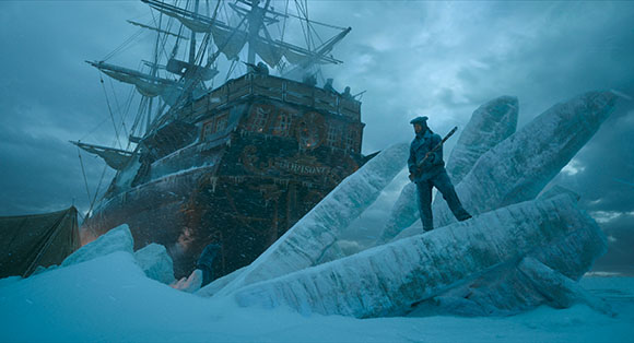 A man in blue clothing stands on large ice formations next to a massive wooden sailing ship in a snowy landscape.