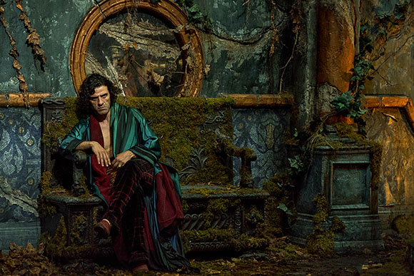 A man in a teal and red robe sits on a mossy stone bench in a dark, decaying room with a large round mirror.