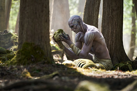A pale, muscular humanoid figure crouches in a sunlit forest, holding a moss-covered object with both hands.