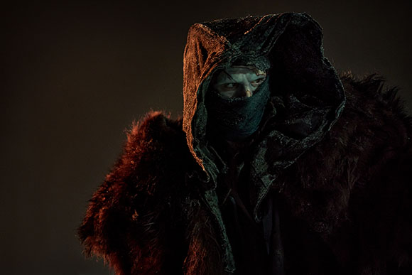 A person wearing a dark, textured hood and a face mask, partially obscured by shadows.