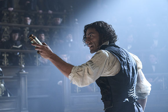 A man with curly hair in a white shirt and dark vest holds a small brass object in a theater.