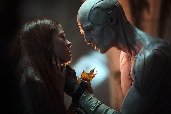A woman with long red hair gazes into the eyes of a pale, muscular humanoid figure holding a single leaf.