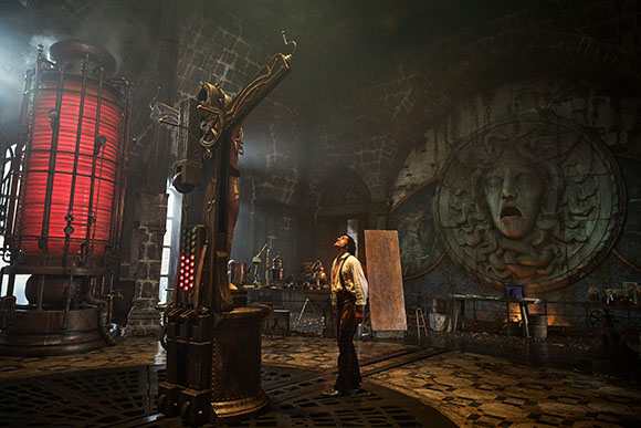 A man stands in a dark, cavernous workshop with large red and metal machinery and a stone face relief.