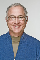 A smiling man with short grey hair and glasses wearing a blue zip-up sweater over a tan turtleneck.