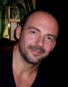 A man with short dark hair and a light beard smiling at the camera against a dark background.