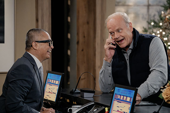 Two men in business attire laugh together across a desk with computer monitors.